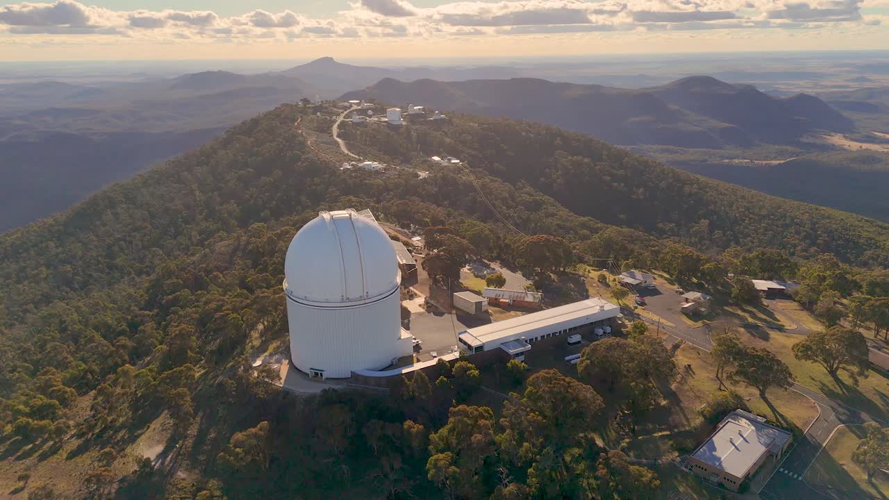 Aerial drone footage circles a large astronomical observatory atop a forested mountain ridge at sunset, revealing surrounding research buildings and expansive landscape views