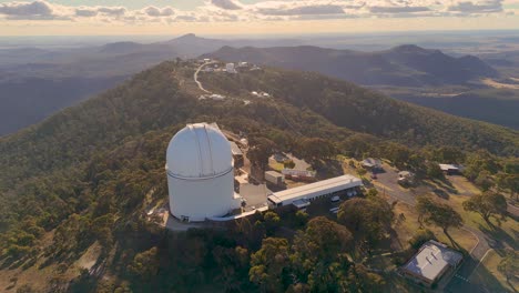 aerial drone footage circles a large astronomical observatory atop a forested mountain ridge at sunset, revealing surrounding research buildings and expansive landscape views
