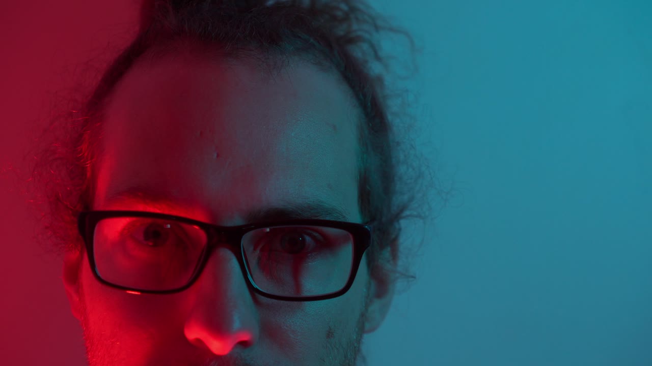 Portrait of a man wearing glasses in moody blue and red lighting, introspective expression as warm red light illuminates half of face