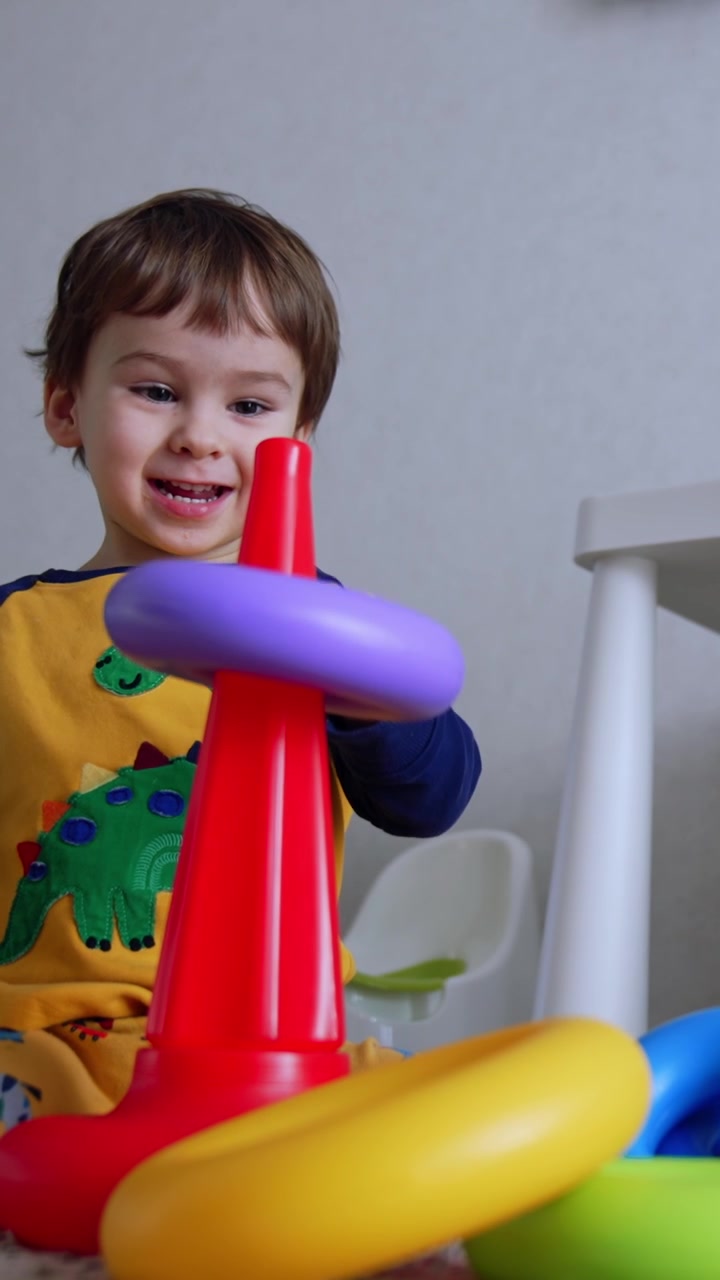 Adorable niño sonriente sentado en el suelo con una pirámide de juguete. Lindo niño jugando felizmente en casa. Vista en contrapicado. Vídeo vertical.