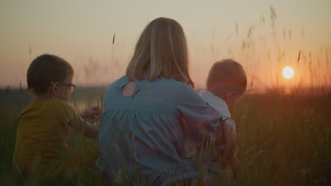 a close-up back shot of a mother in a blue gown sitting in a field during sunset, holding her younger child on her lap while the older child sits beside her. they all gaze out at the serene lake