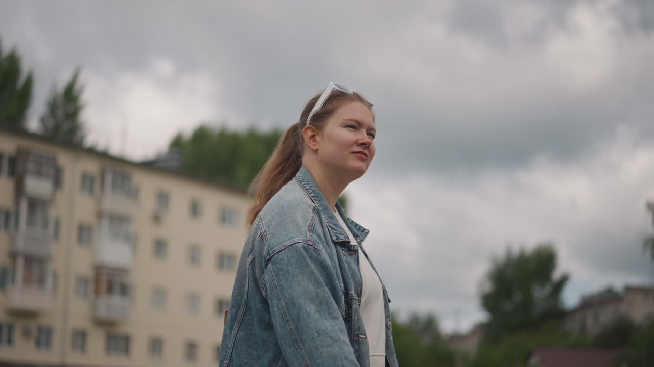 Hvid kvinde står under dramatiske skyer, denimjakke og hårbånd mod en lejlighedsbygning, overskyet himmel, eftertænksomt blik, studerende holder pause under gåtur i nabolaget, introspektivt filmisk.