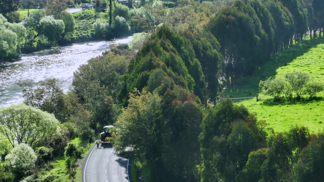 Drone shot capturing a tractor driving down a road past lush green fields on a sunny day in New Zealand.