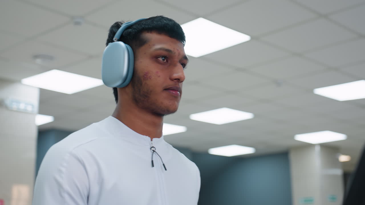 young man clips over ear headset onto neck steps onto treadmill powers console and begins cardio session in spacious gym lit by overhead panels with equipment and exerciser blurred in background
