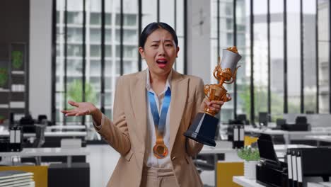 Wondering Asian Business Woman In A Suit With A Gold Medal Looking At Trophy Doubtfully In The Office