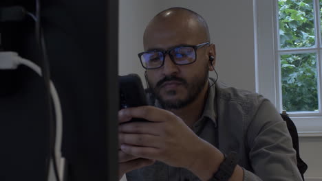modern uk asian male wearing eyeglasses in office using mobile phone at desk. locked off