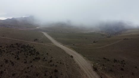 Low-cloud-aerial-follows-gravel-road-on-foggy-mountain-plateau,-Andes