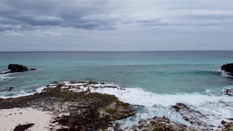 drone aéreo volando desde una playa rocosa y de arena blanca hacia un mar caribeño agitado pero claro en cozumel, méxico