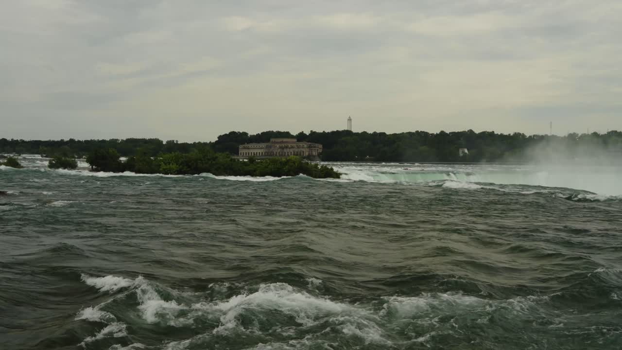 A powerful view of the upper Niagara River just before it plummets over the falls, with an abandoned power plant in the distance and lush greenery bordering the rushing water