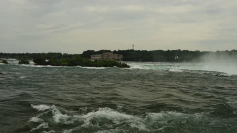 a powerful view of the upper niagara river just before it plummets over the falls, with an abandoned power plant in the distance and lush greenery bordering the rushing water