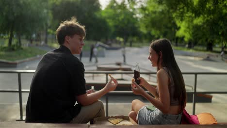 two young friends enjoy pizza and soda while sitting on a skatepark railing