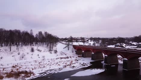 Aerial-view-of-old-red-brick-bridge-across-the-Venta-river-in-Kuldiga,-Latvia-in-overcast-winter-day,-flyover-over-the-trees,-wide-angle-ascending-drone-shot-moving-forward