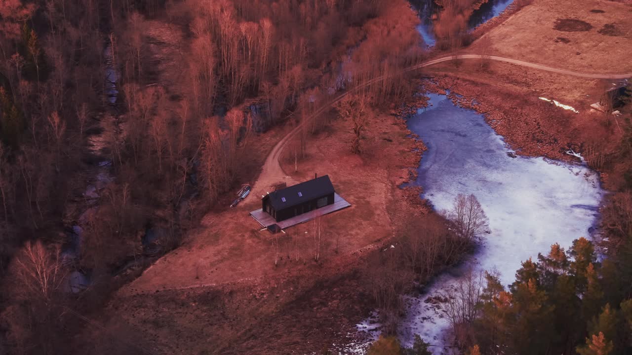 Dronebilde av en svart hytte i en vårskog med en delvis frossen innsjø i bakgrunnen. Kinematisk naturlandskap, ideelt for reise, turisme og bakgrunnsvideo