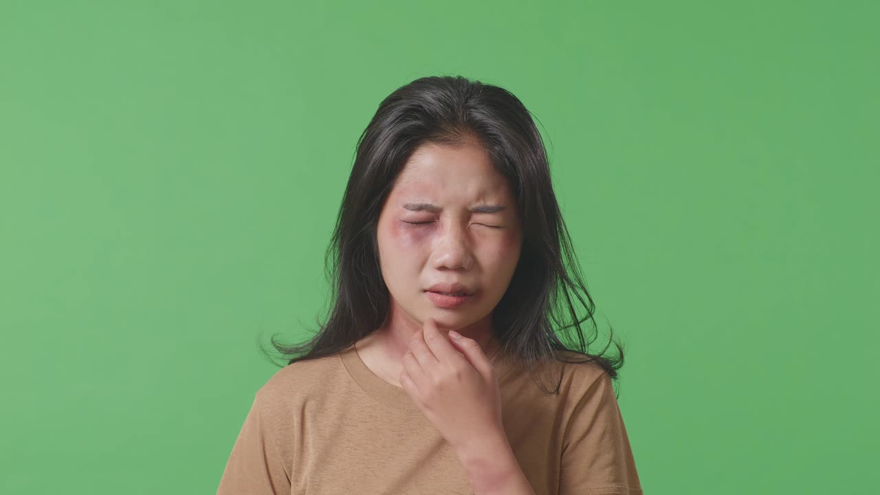 얼굴에 이 있는 젊은 아시아 여성의 폭력 피해자, 그녀의 얼굴을 만지는, 녹색 스크린 배경 스튜디오에서 신체적으로 학대당하는 것에서 상처를 입는.