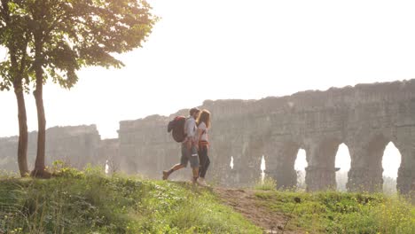 young lovely couple backpackers tourists walking holding hands arrive on hill in front of roman aqueduct arches in parco degli acquedotti park ruins in rome on romantic misty sunrise with guitar and sleeping bag slow motion