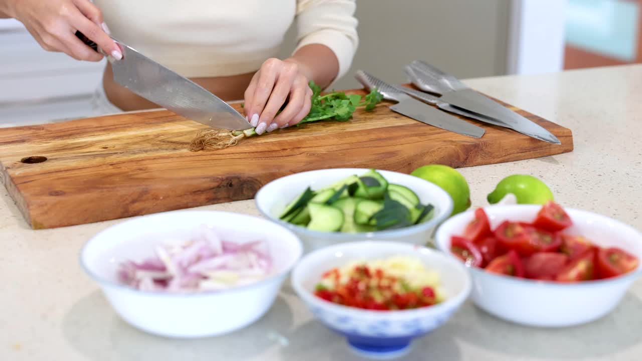 A person chops coriander on a wooden board surrounded by fresh vegetables in a well-lit kitchen