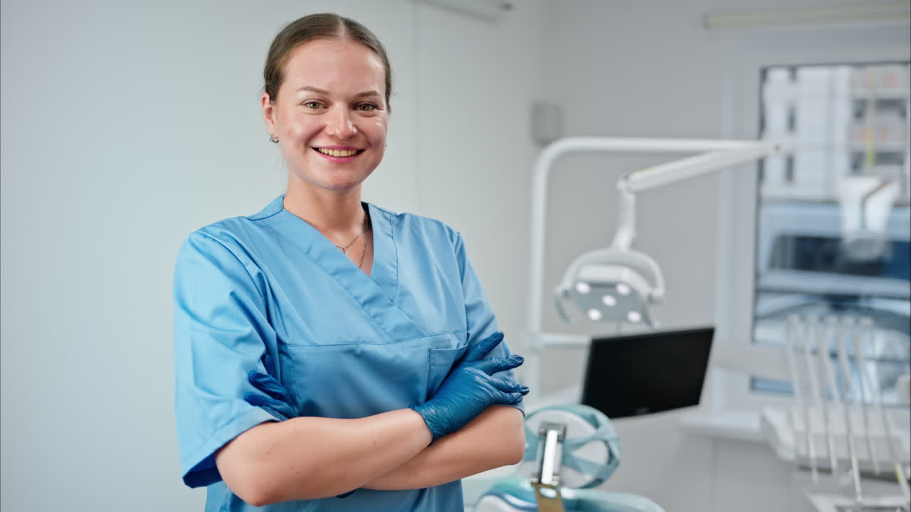 Dentist wearing a scrub and standing and smiling in a dental cabinet, in front of a window