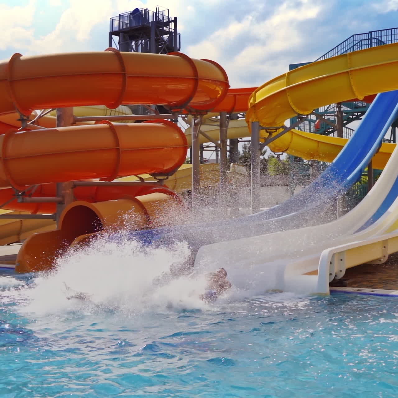 People ride down from colorful water slides. Beautiful water splash in the outdoor pool. Family entertainment in summer. Slow motion.