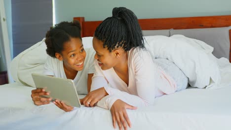 Vorderansicht-Einer-Mittleren-Erwachsenen-Schwarzen-Mutter-Und-Tochter,-Die-Ein-Digitales-Tablet-Auf-Dem-Bett-In-Einem-Komfortablen-Zuhause-In-4K-Nutzt