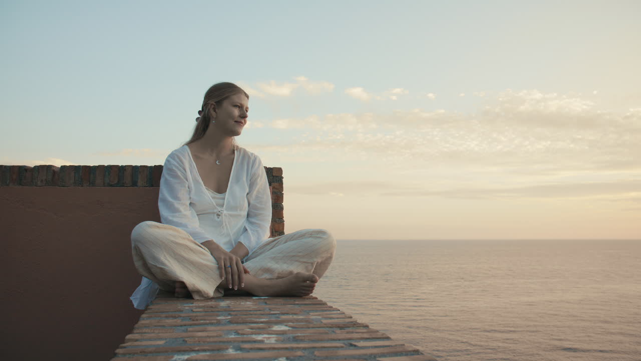 Wide static shot a showing a serene woman sitting cross-legged on a brick ledge, bathed in soft morning or evening light—open, tranquil, reflective. Watching the ocean