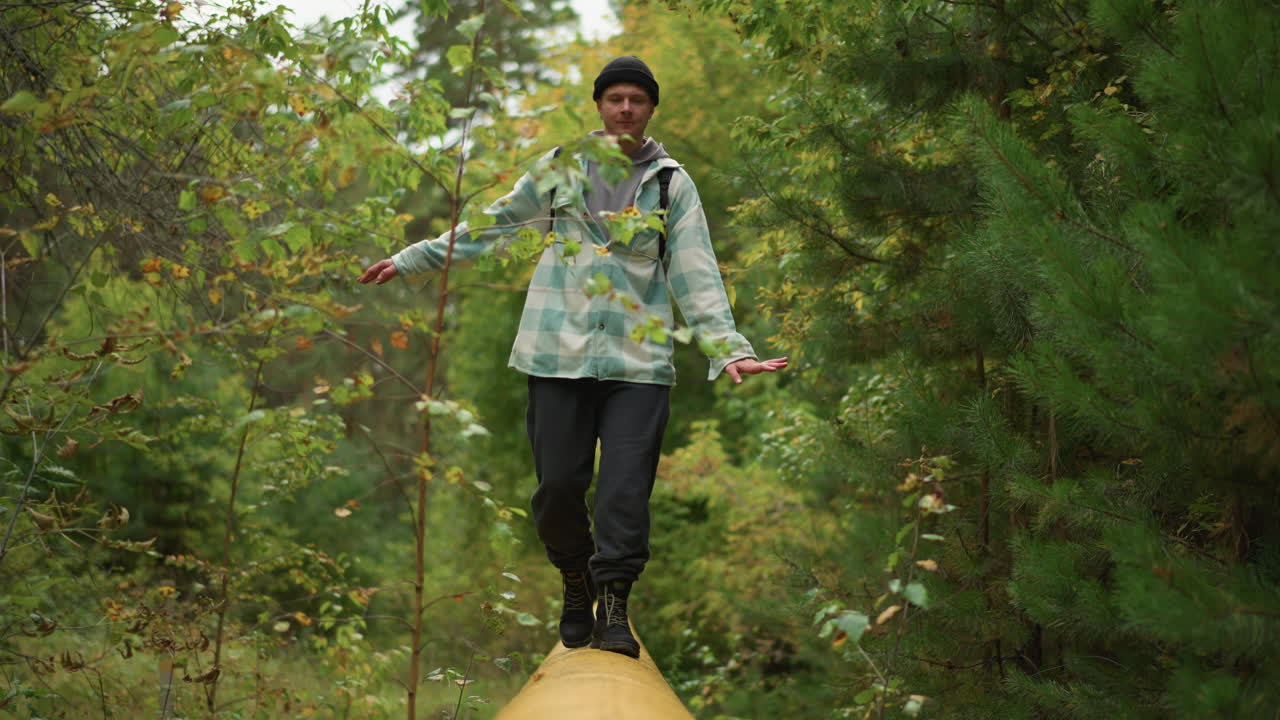 Caucasian explorer pausing to admire canopy, looking upward while balancing on yellow pipe amid lush green trees, contemplative expression, backpack and casual outdoor gear, gentle sunlight filtering