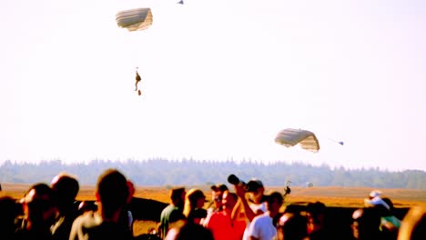 multiple parachutists are landing in a wide, open field during daylight. a crowd is visible at the bottom, watching the event unfold. the clear sky and expansive landscape provide a serene atmosphere