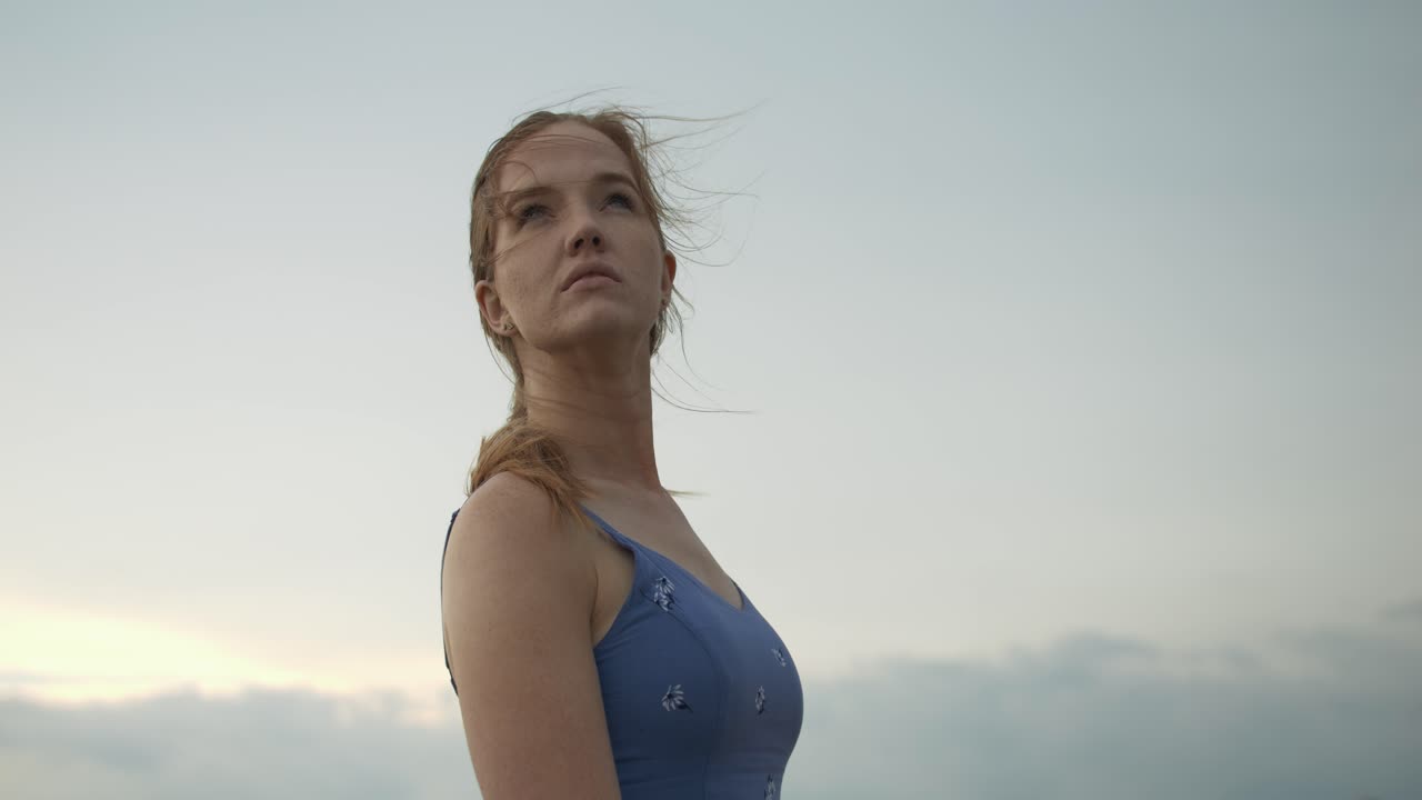 A young woman stands in the middle of a wheat field wearing a light blue dress with flowers, swaying gently in the breeze under a dramatic, stormy sky. The scene captures the essence of rural life.