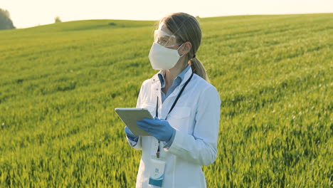 caucasian researcher woman in white coa, mask and goggles using tablet and looking around in the green field