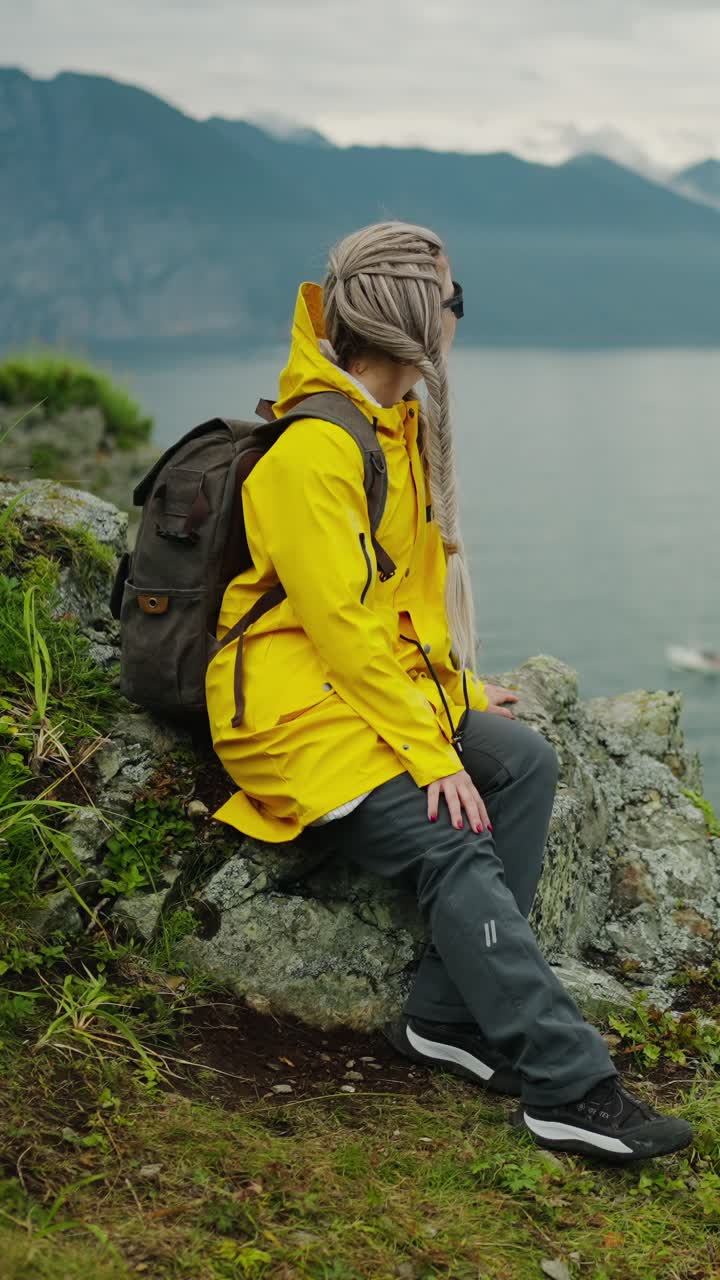Escursionista donna con impermeabile giallo seduta su una scogliera di montagna con vista lago