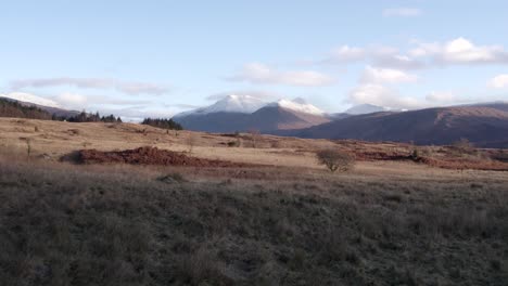 aerial drone footage flying low and close to bracken and moorland towards glen etive and loch etive in the highlands of scotland with snow-capped mountains, a forest and still water in morning light