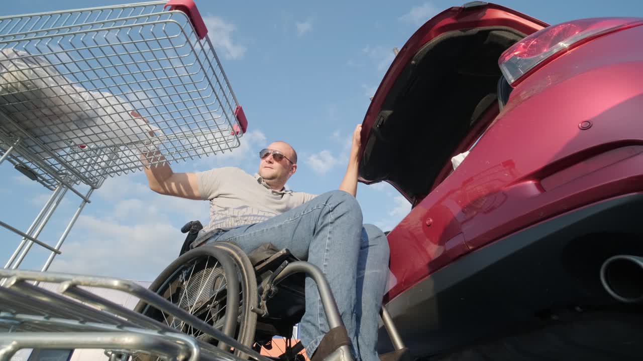 체어에 앉은 장애인 성인 남자가 슈퍼마켓 주차장에서 차의 트크에 물건을 넣고 있다.