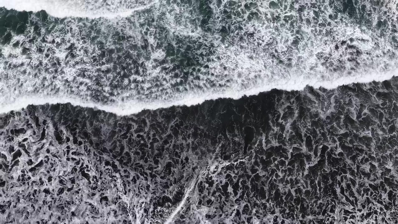 Drone view of white foam waves washing onto black volcanic sand beach from above, New Zealand North Island, moody nature and dramatic coastal view.