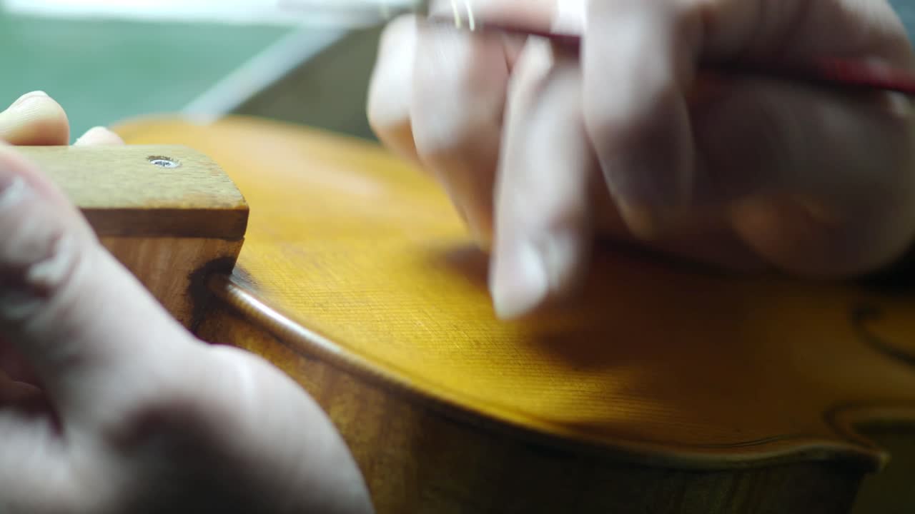 Liutaio concentrato sull'antichizzazione del filetto su un violino di nuova produzione, con tecnica a dito e pennello, che mostra una delicata tecnica di lavorazione del legno e la maestria nello strumento musicale.