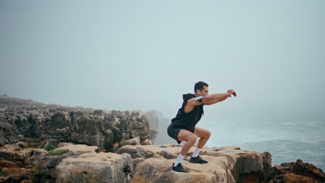 fit athlete doing squats on seaside cliff. focused sporty guy training outdoors