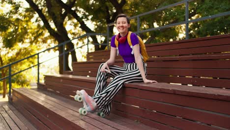 A-girl-with-a-short-haircut-in-a-purple-top,-striped-pants-and-a-yellow-backpack-sits-on-brown-benches-in-the-stands-in-pink-roller-skates-in-a-skatepark-in-summer