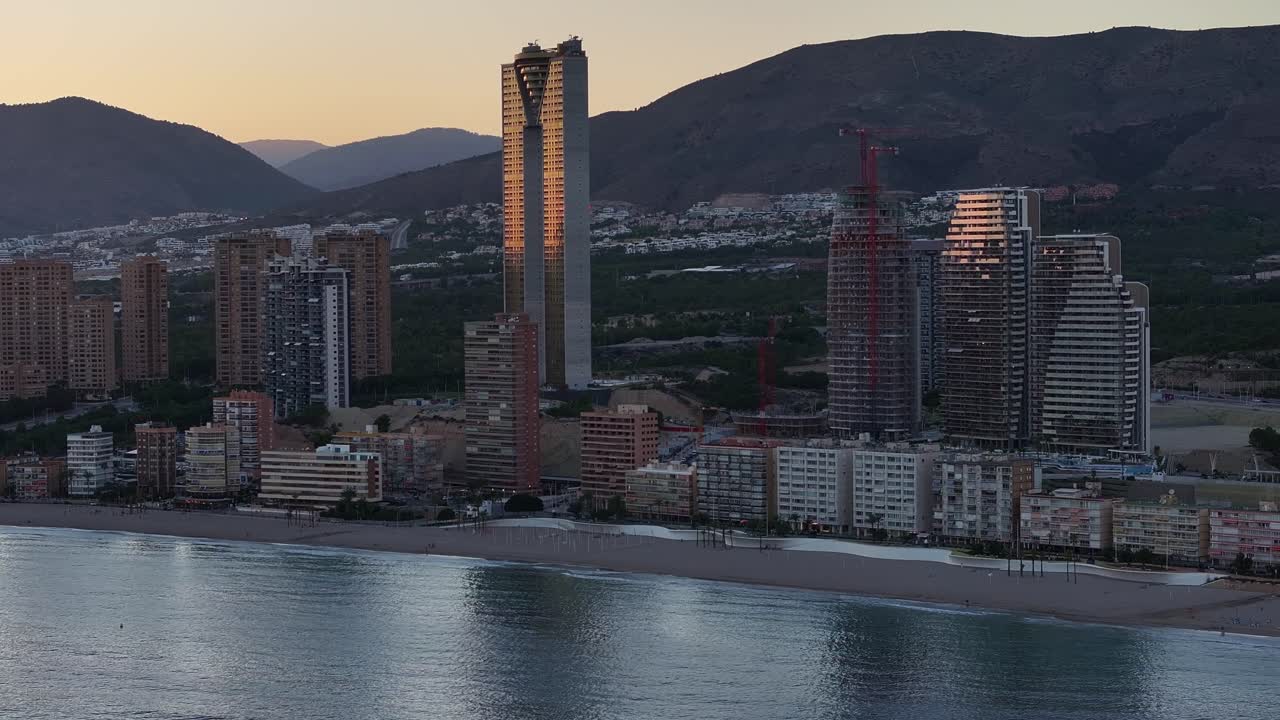 Il drone si muove lateralmente lungo il tranquillo lungomare di Benidorm all'alba, mostrando la spiaggia, alti edifici con riflessi del sole, montagne e traffico leggero.