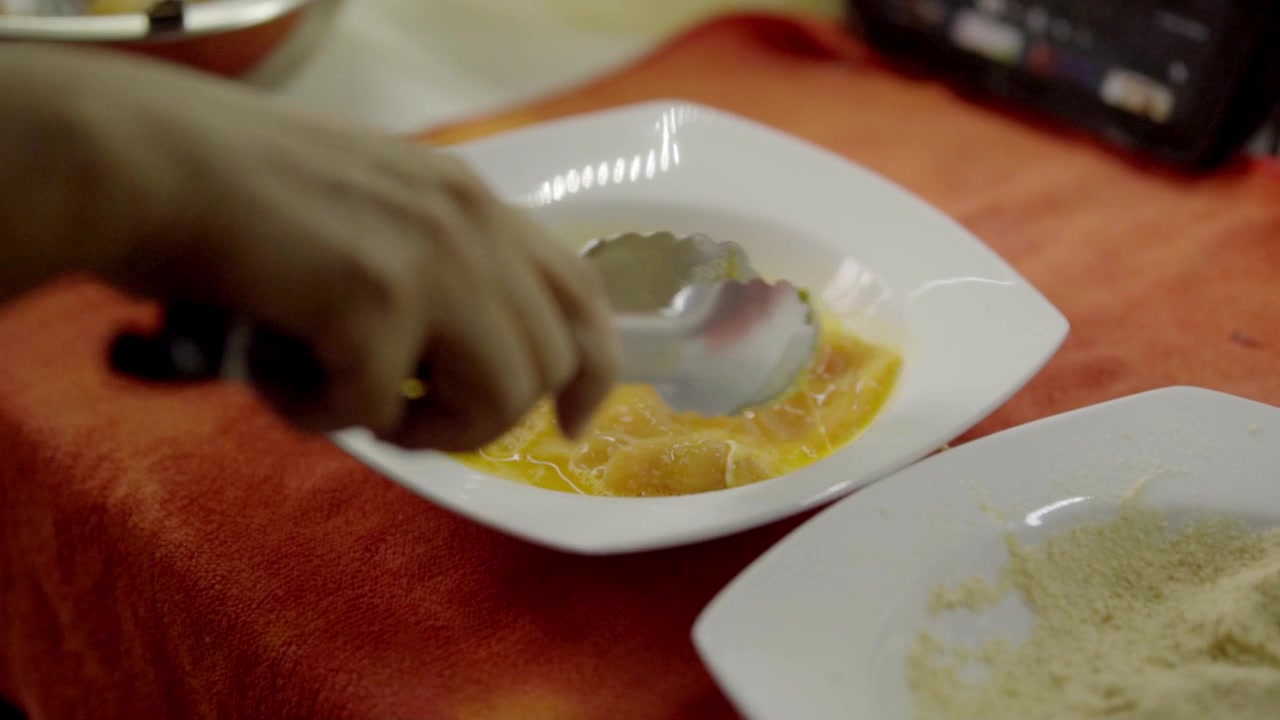 Vista de cerca de una mano cubriendo trozos de pollo en mezcla de huevo dentro de un tazón blanco, plato de harina cerca, paso del proceso de empanado para cocinar, escena sencilla de preparación de alimentos en la cocina.