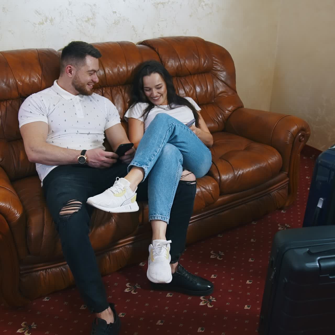 Happy young couple on a sofa in hotel. Smiling man and woman in casual clothes sitting together with mobile phones and talk on suitcases background