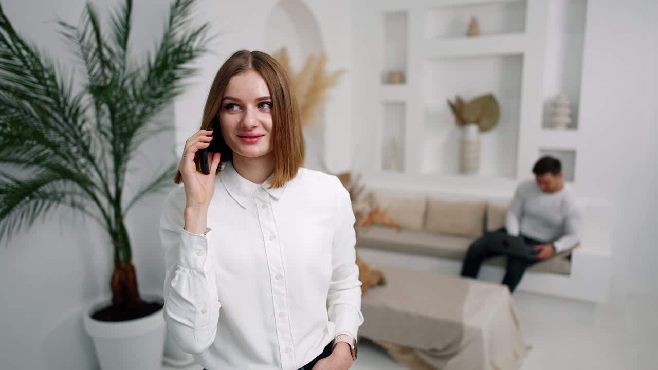 Caucasian woman speaking on the phone looking at camera. Man sitting at backdrop in blur uses laptop.