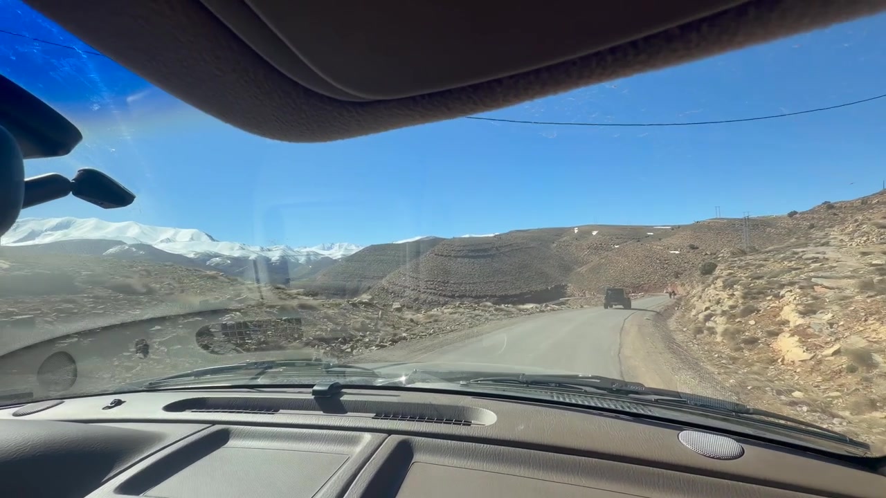 POV dall'interno di un'auto che guida lungo una strada di montagna nelle montagne dell'Atlante in Marocco, con terreno roccioso e cime innevate in lontananza.