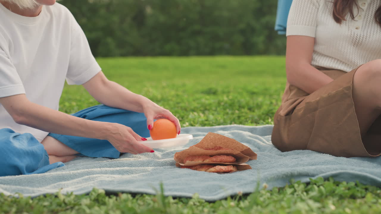 lower view of young mothers sharing fruit on picnic blanket with sandwich, one handing orange to other, relaxed summer park scene, hands and legs visible on soft towel, green grass background