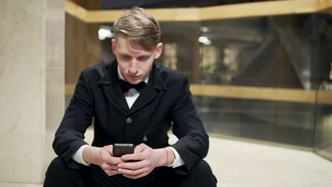 Young-blonde-man-in-black-suit-with-smartphone-sitting-on-stairs-in-hotel