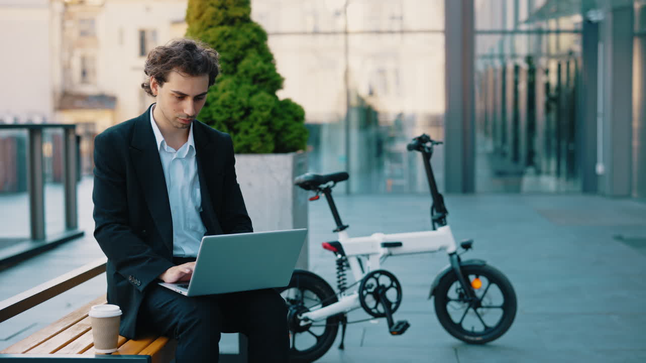 Man working on laptop on a bench with an electric bike nearby in a city setting