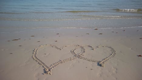 static shot of two interlocking hearts on a beach with water on lapping up on the beach