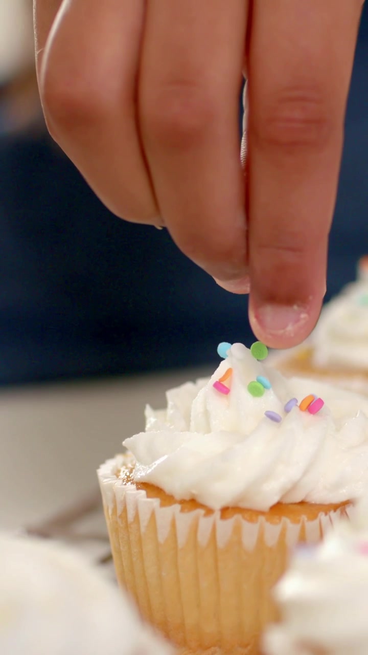 Vídeo vertical: Mão de mulher com manga pegando um granulado e colocando-o em um cupcake no balcão, decorando.