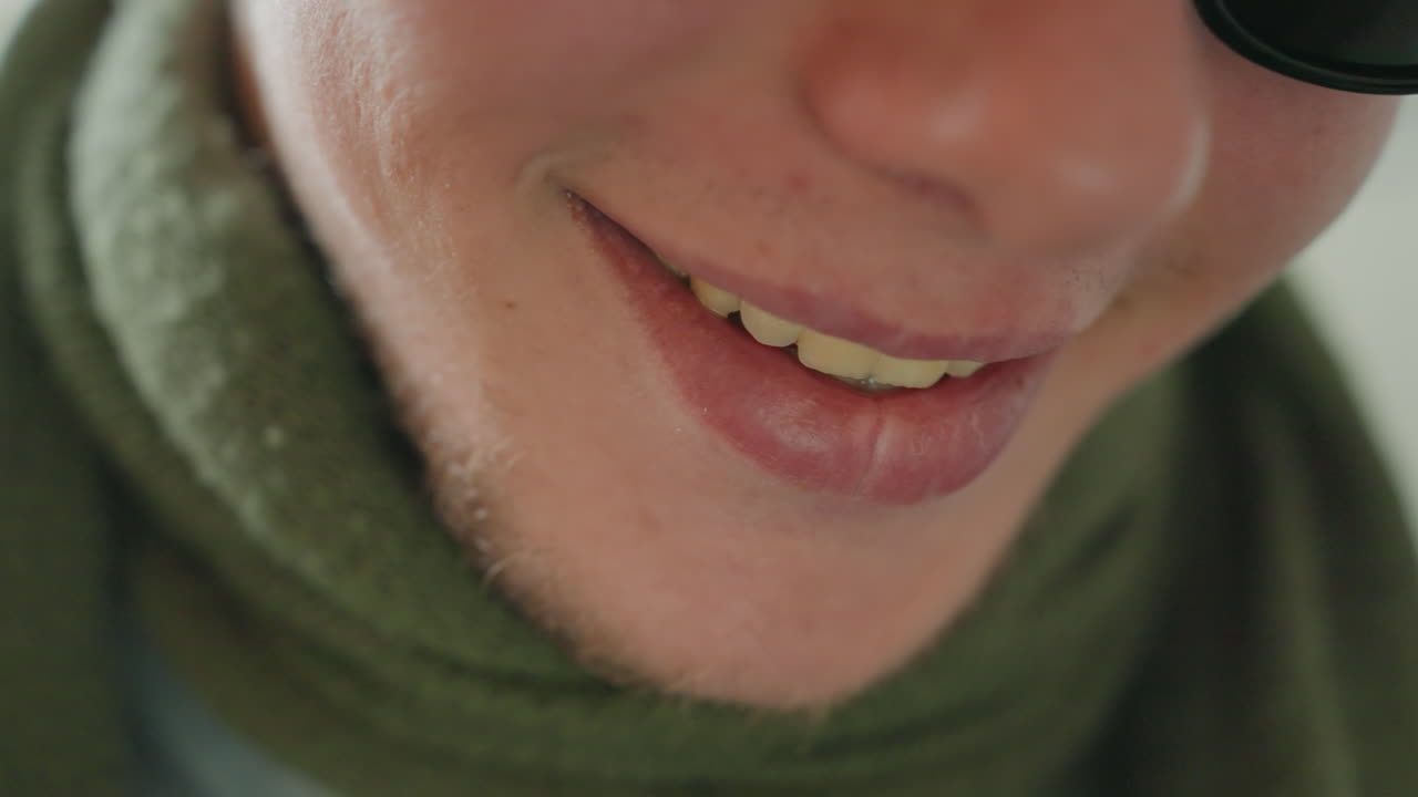 Homem branco sorrindo, exibindo um riso caloroso, com cachecol verde e óculos de sol em close-up. Um sorriso atrevido revelando os dentes, transmitindo uma vibe alegre e espontânea. Luz interna aconchegante e energia de narrador brincalhão e acessível.