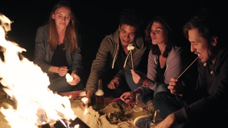 Young-cheerful-friends-sitting-by-the-fire-on-the-beach-late-at-night,-roasting-marshmallow-on-sticks-over-the-fire-together