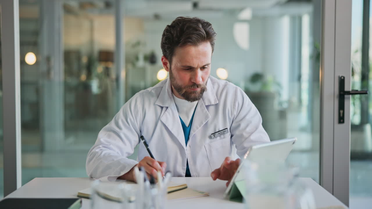 Male Doctor Writing Notes and Using Tablet at Desk