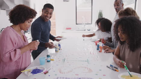 Familia-Negra-De-Tres-Generaciones-Preparando-Un-Cartel-Para-Una-Fiesta-Sorpresa-En-Su-Cocina,-Tiro-Inclinado