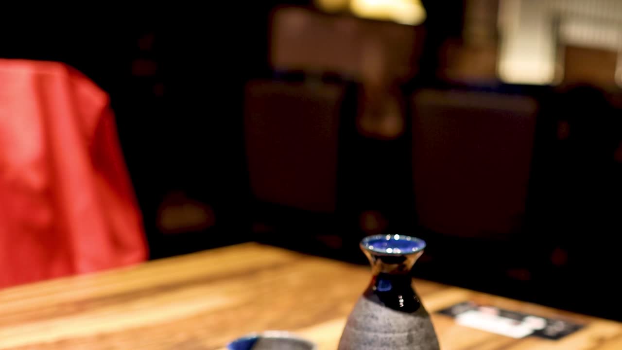 A hand grasps a frothy beer mug beside a traditional sake bottle on a wooden table.